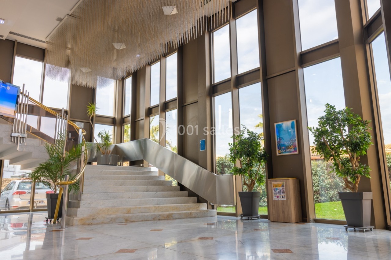 Hall lumineux avec escalier en marbre, grandes baies vitrées, plantes décoratives et vue sur l'extérieur.