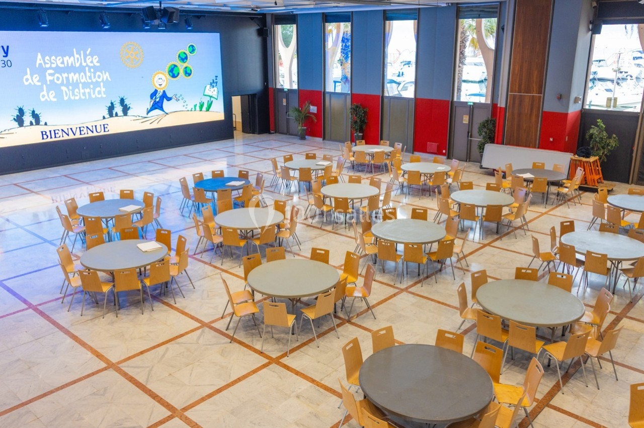 Salle de conférence avec des tables rondes et des chaises disposées, écran de présentation en arrière-plan.