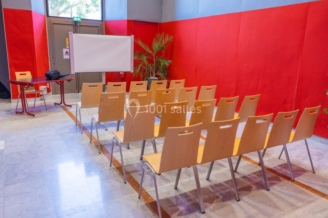 Salle de réunion avec des chaises en bois alignées face à un écran blanc, entourée de murs rouges.