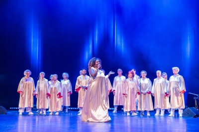 Une chanteuse en robe dorée se produit sur scène, entourée d'un chœur en tenues assorties, sous un éclairage bleu.