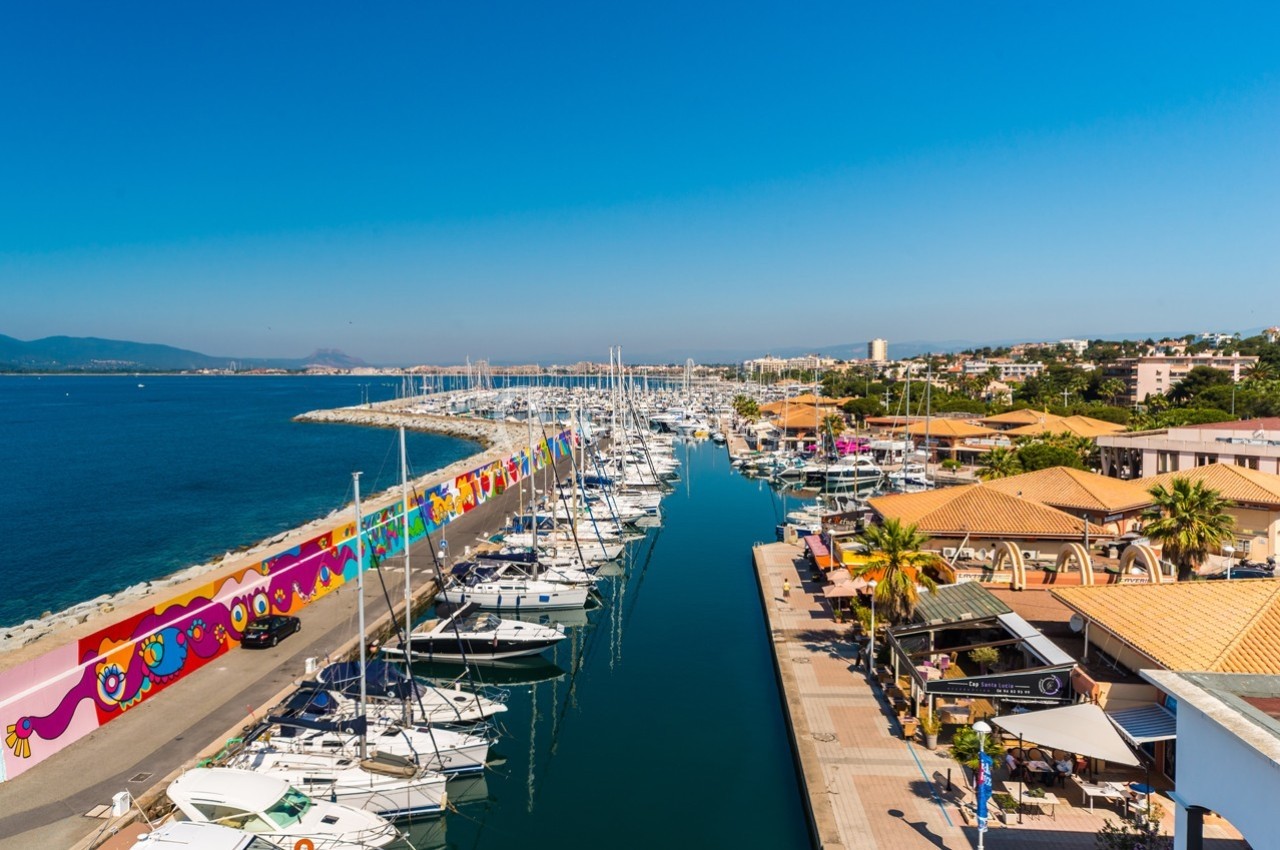 Vue d'un port de plaisance avec des bateaux amarrés, une promenade bordée de bâtiments et une fresque colorée sur un mur.