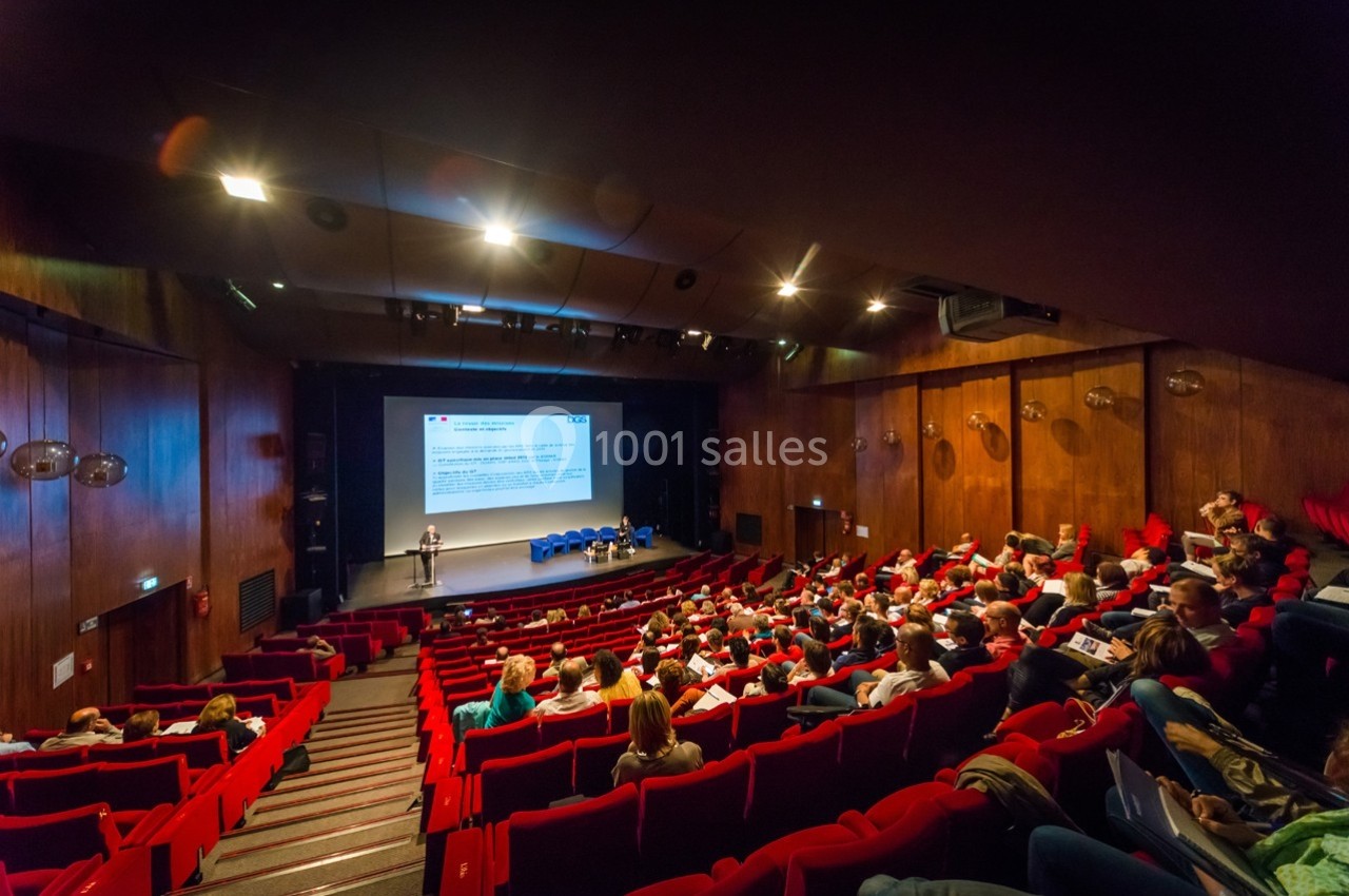 Salle de conférence avec des participants assis face à une scène où une personne présente un diaporama.