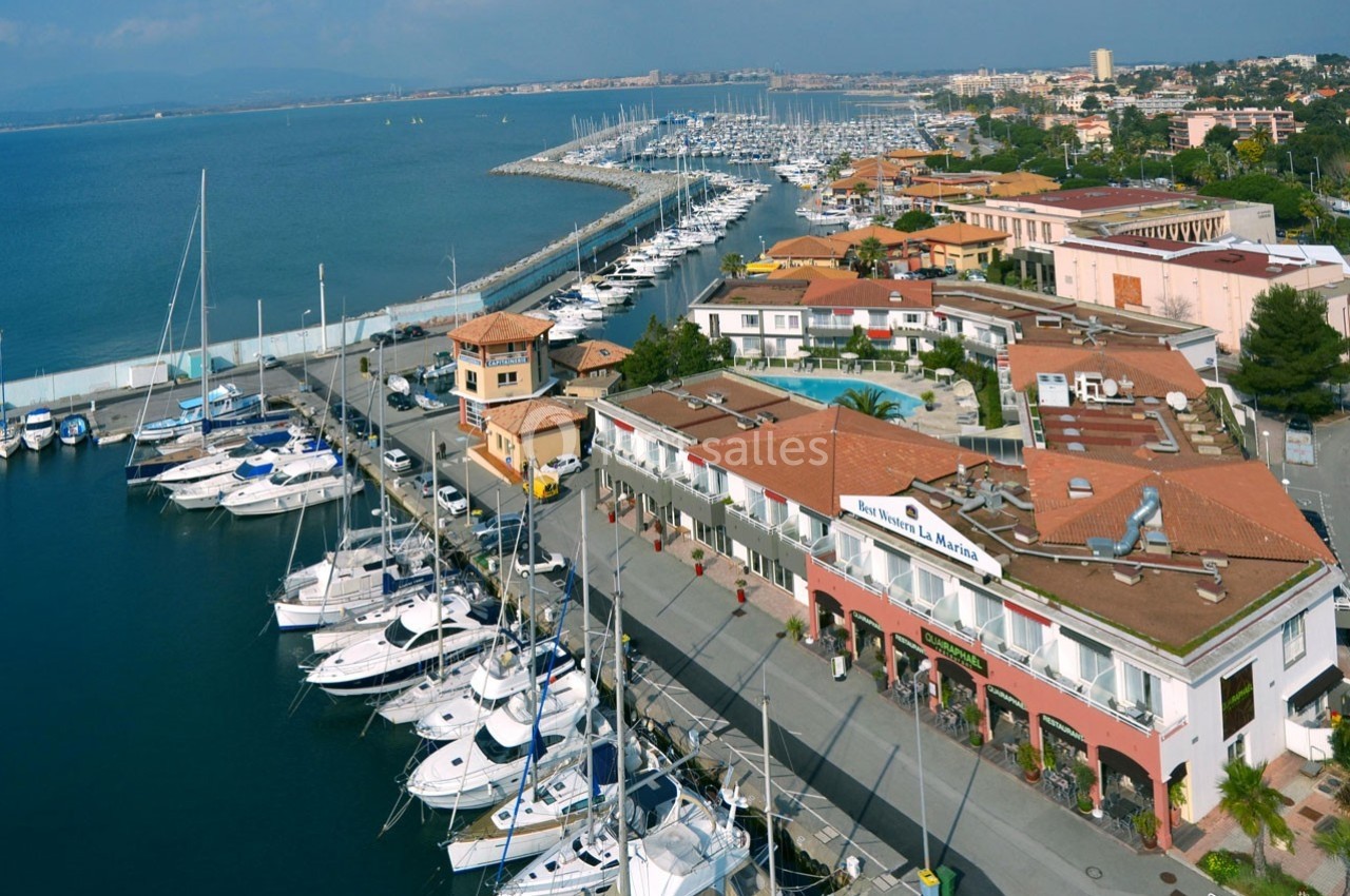 Vue aérienne d'un port de plaisance avec des voiliers amarrés, des bâtiments commerciaux et une promenade en bord de mer.