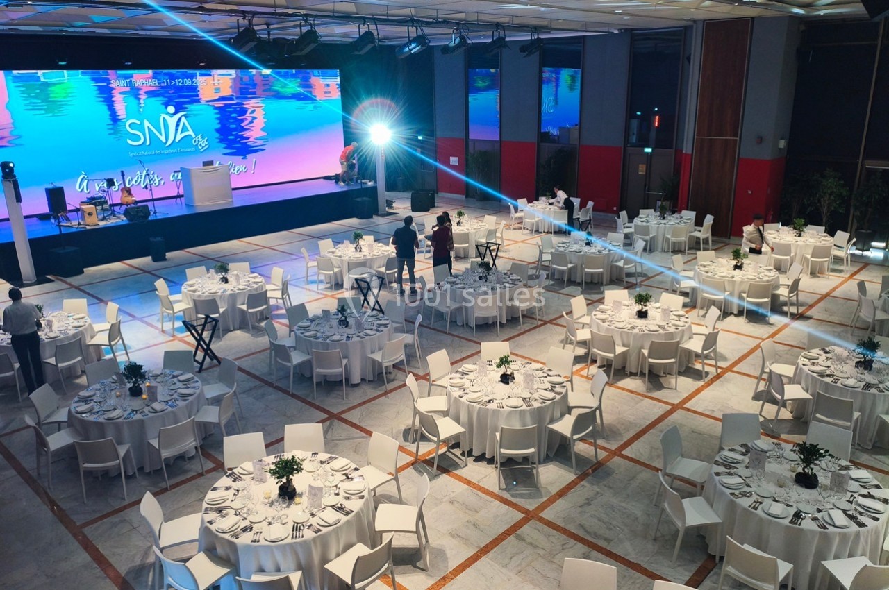 Salle de réception avec tables rondes dressées, éclairage lumineux et écran géant en arrière-plan.