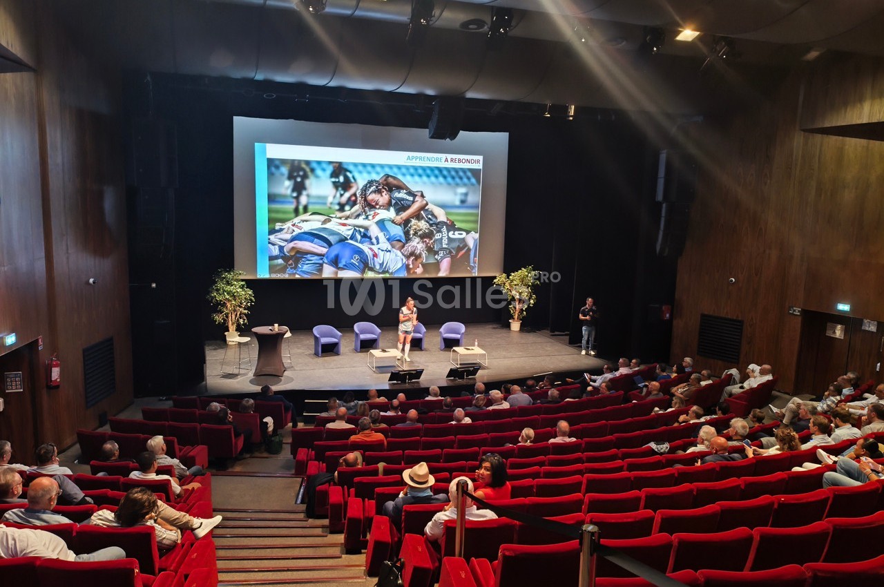 Conférence dans un auditorium avec une présentation projetée sur scène et un public assis dans des fauteuils rouges.