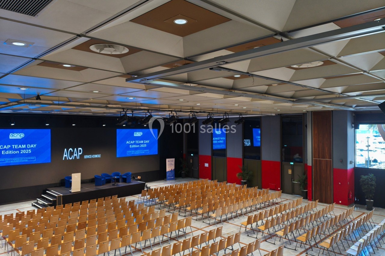Salle de conférence moderne avec des rangées de chaises vides, une scène équipée d'écrans et un podium.