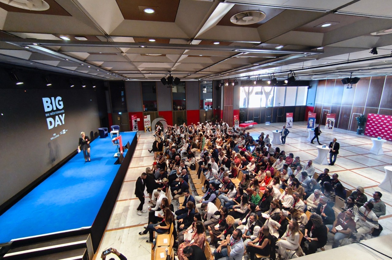Salle de conférence remplie de participants, avec une scène bleue où deux intervenants s'expriment devant un écran.