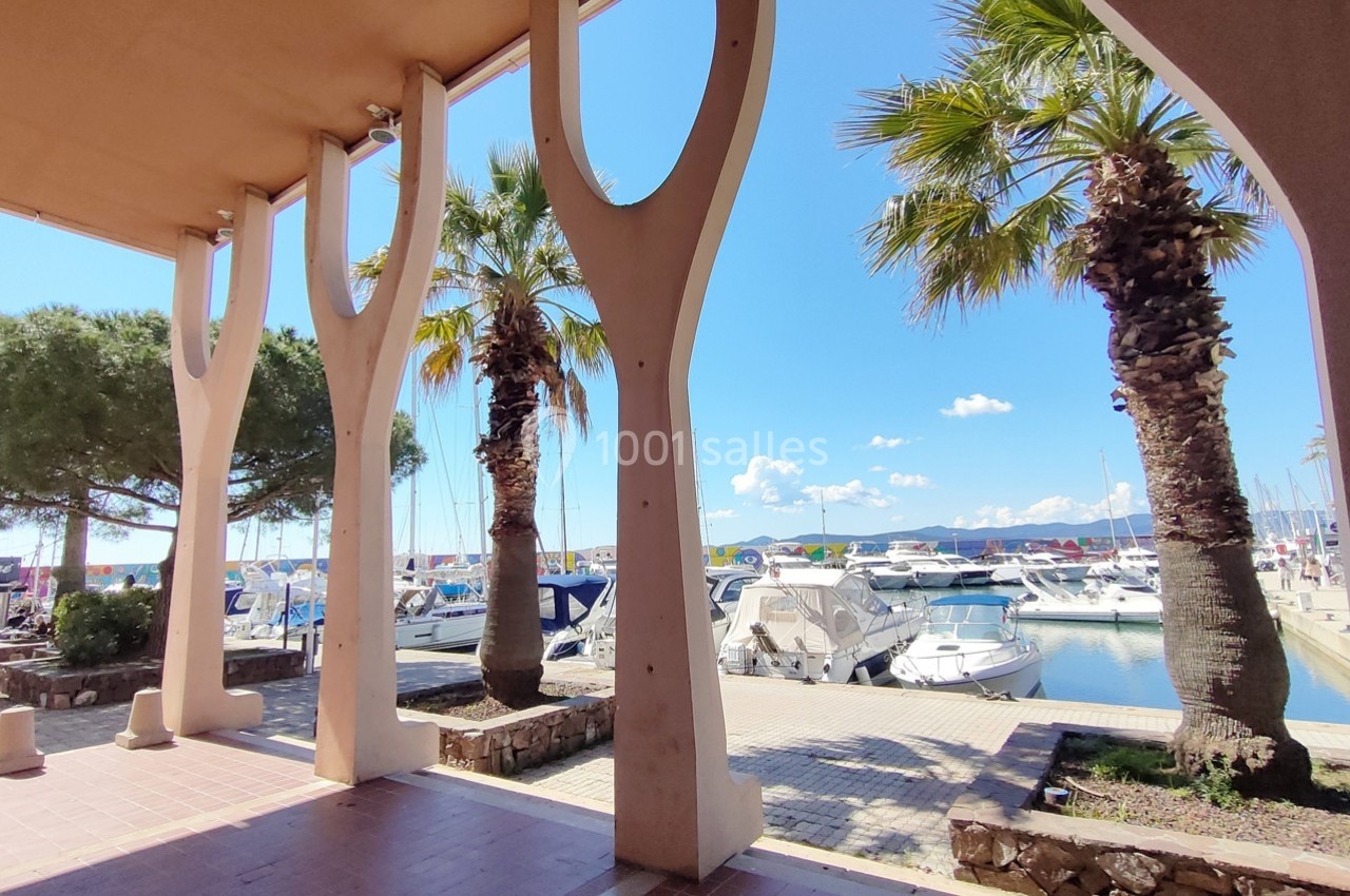 Vue sur un port de plaisance avec des bateaux amarrés, encadrée par des colonnes et des palmiers sous un ciel bleu.