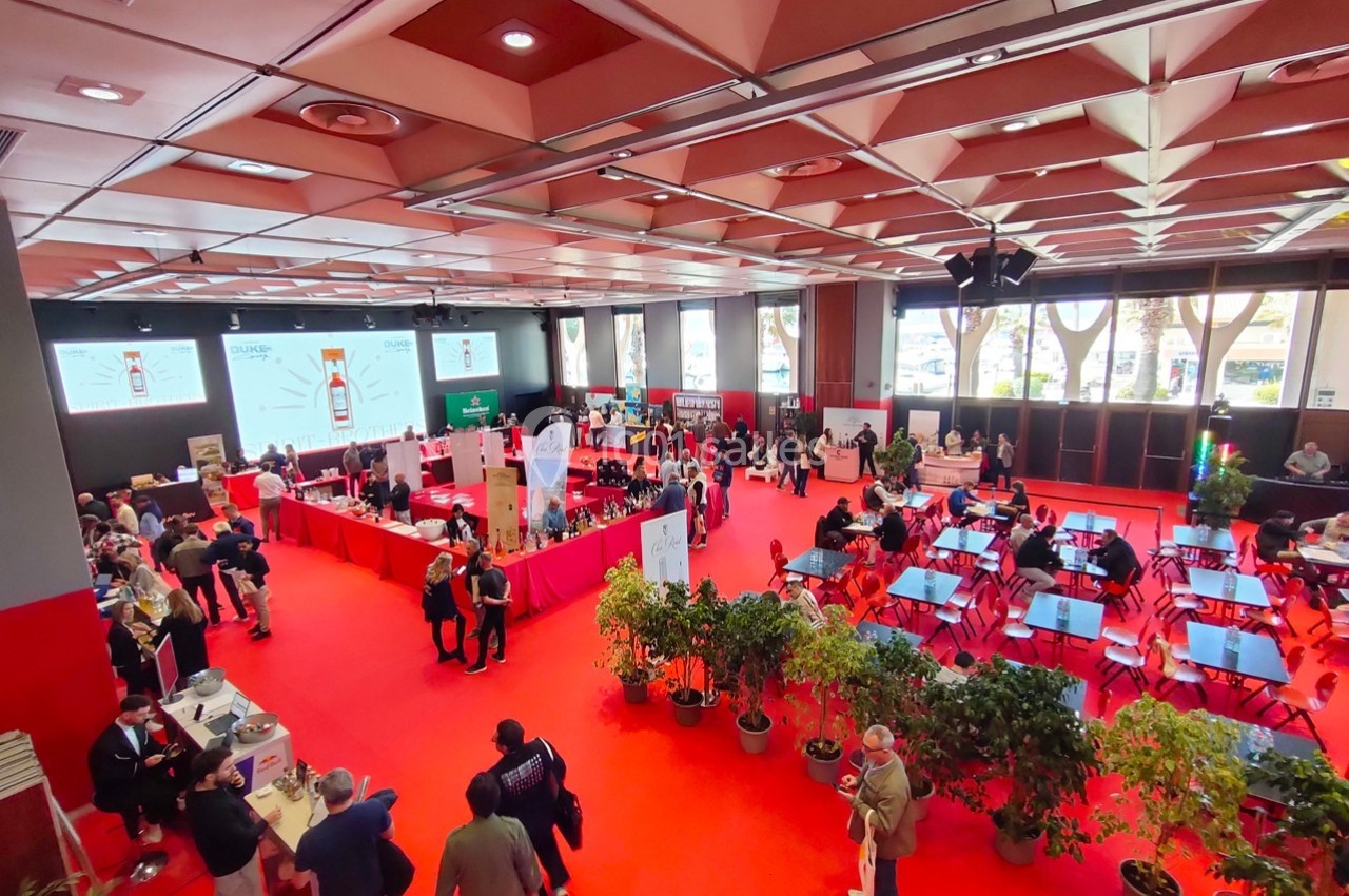 Salle d'exposition animée avec des stands, des visiteurs et des plantes décoratives sur un sol rouge.