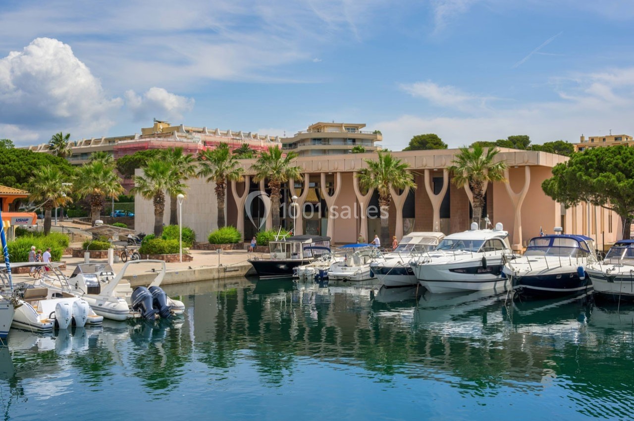 Port avec des bateaux amarrés devant un bâtiment moderne entouré de palmiers, sous un ciel partiellement nuageux.