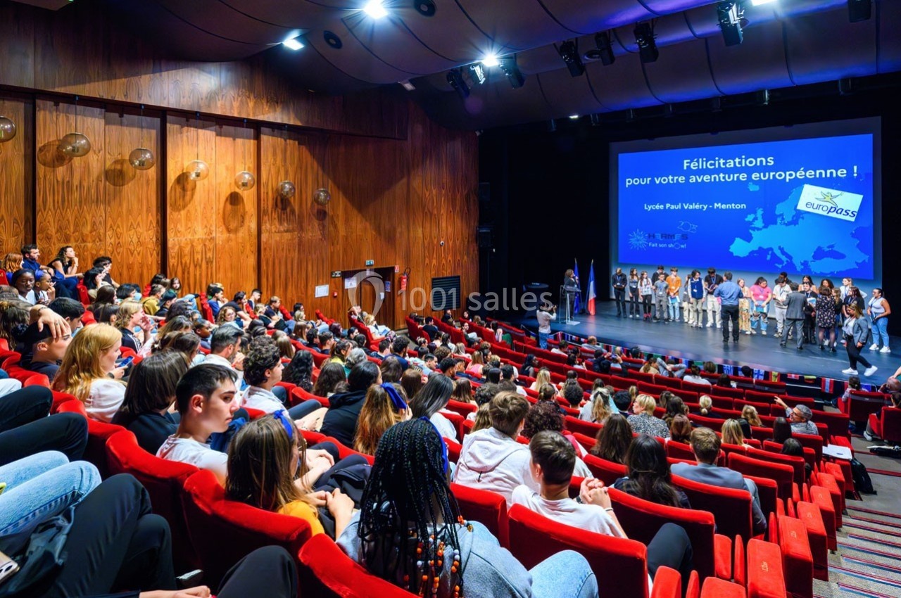 Un amphithéâtre rempli de spectateurs face à une scène où des personnes sont alignées devant un écran de projection.