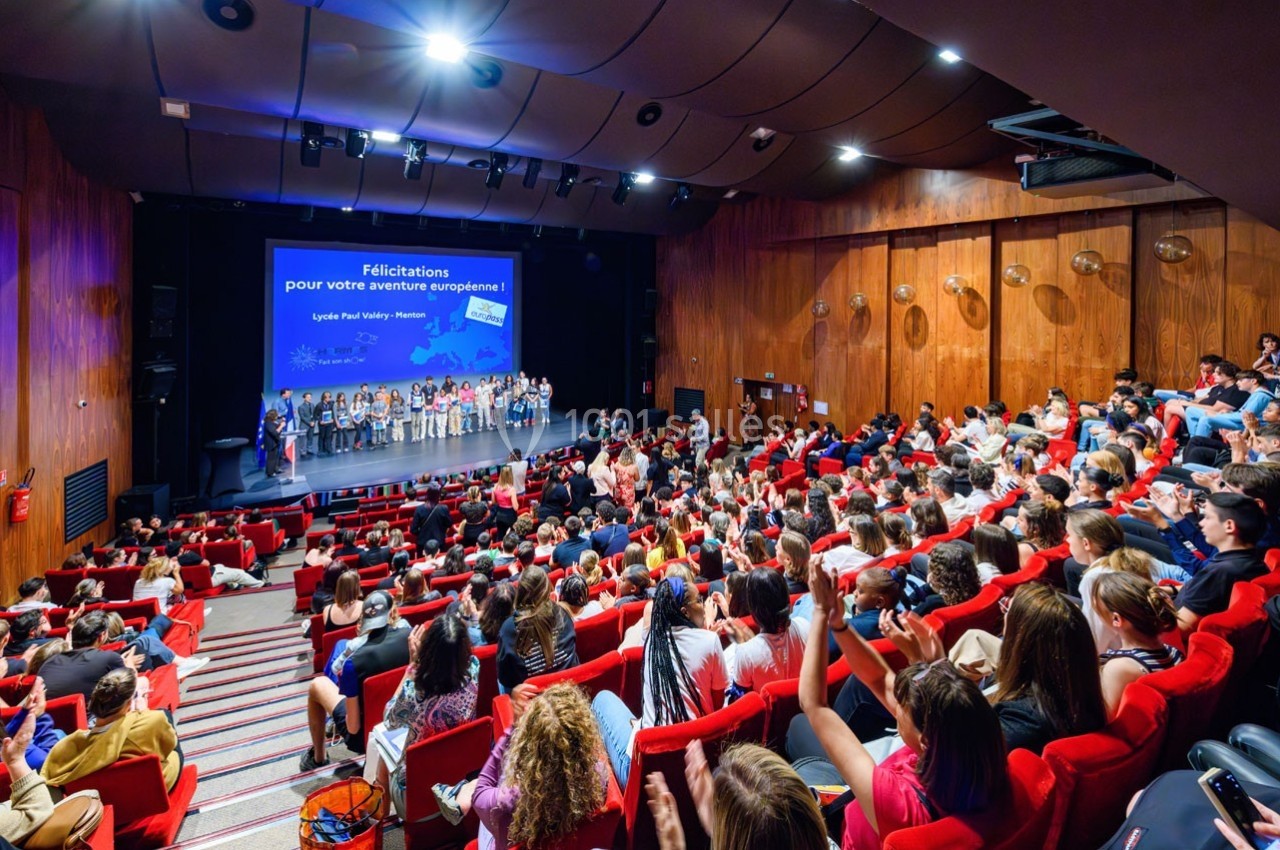 Salle comble avec des spectateurs assis face à une scène où des personnes sont réunies devant un écran de projection.