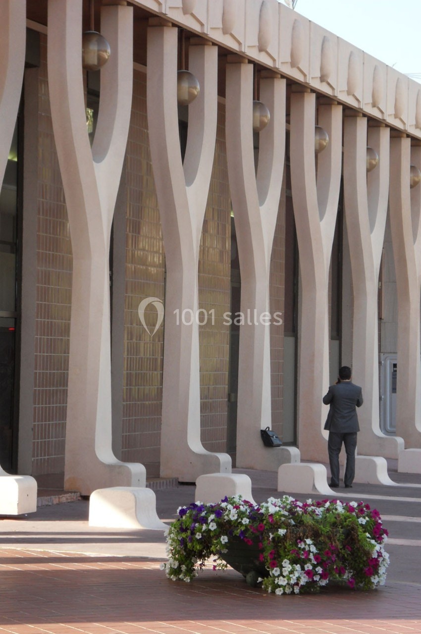Façade moderne avec colonnes en forme d'arbre, un homme en costume debout et un parterre de fleurs colorées au premier plan.