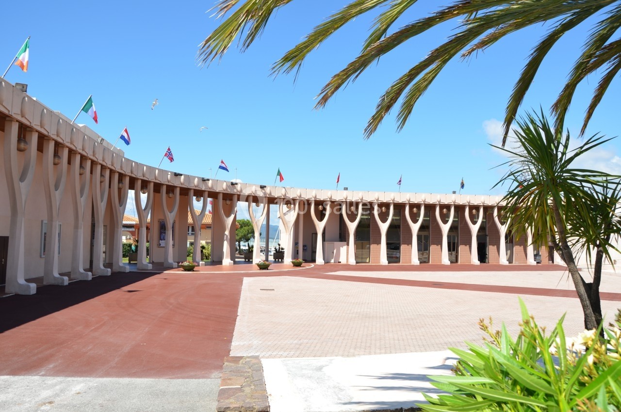 Bâtiment avec une architecture en arcades, entouré de drapeaux internationaux et de palmiers sous un ciel bleu.