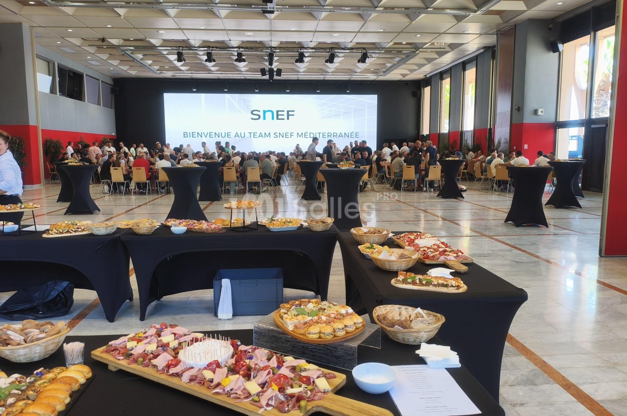 Salle de congrès aménagée pour un événement - tables hautes noires avec mets, gradins de convives, écran de projection et plafond lumineux.