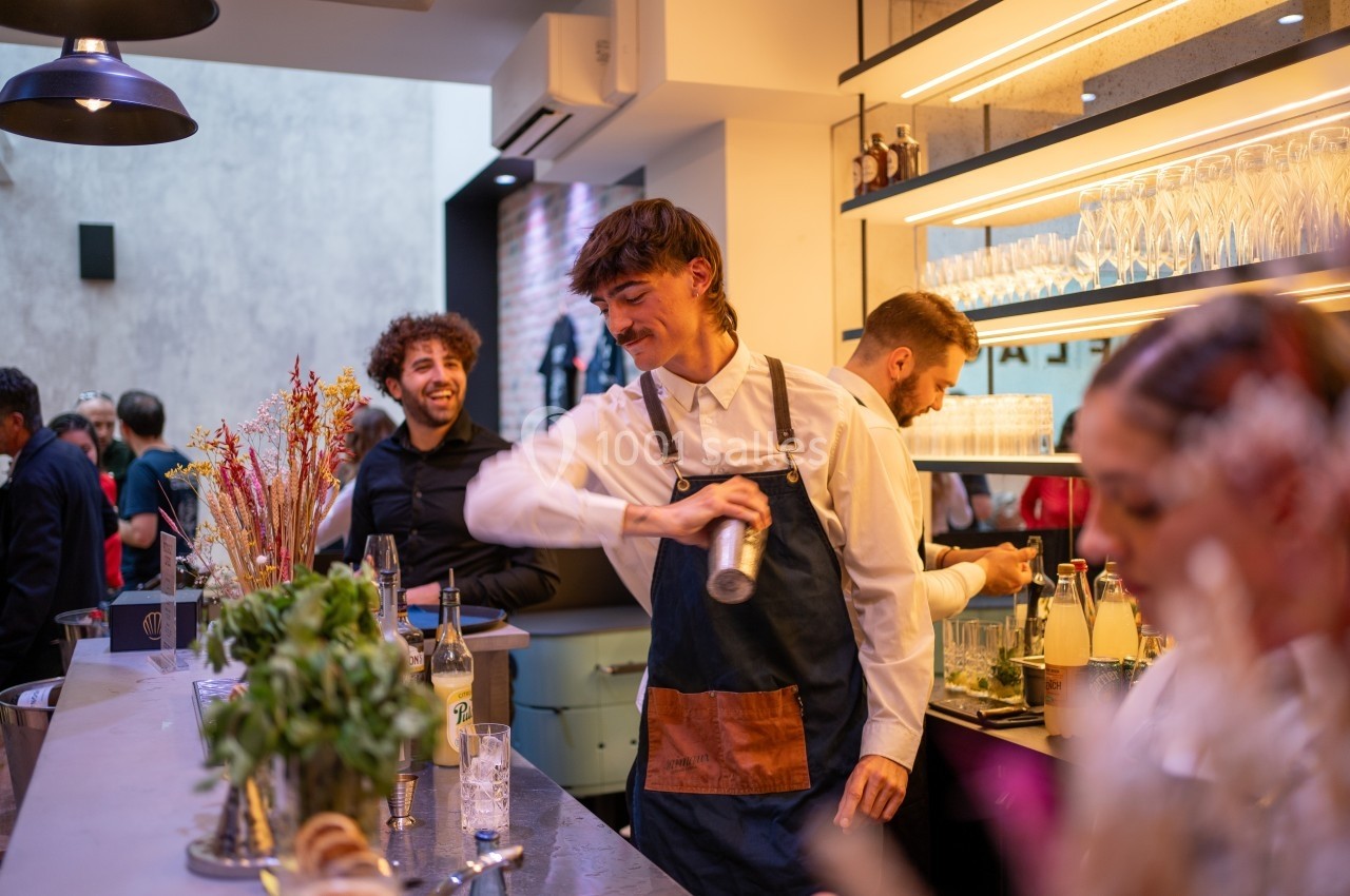 Un barman prépare un cocktail avec un shaker derrière un comptoir, entouré de clients dans un bar animé.