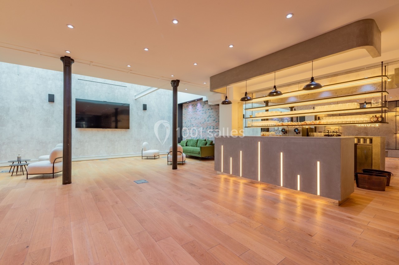 Espace intérieur moderne avec parquet en bois, bar éclairé, canapé vert et murs en béton et briques apparentes.