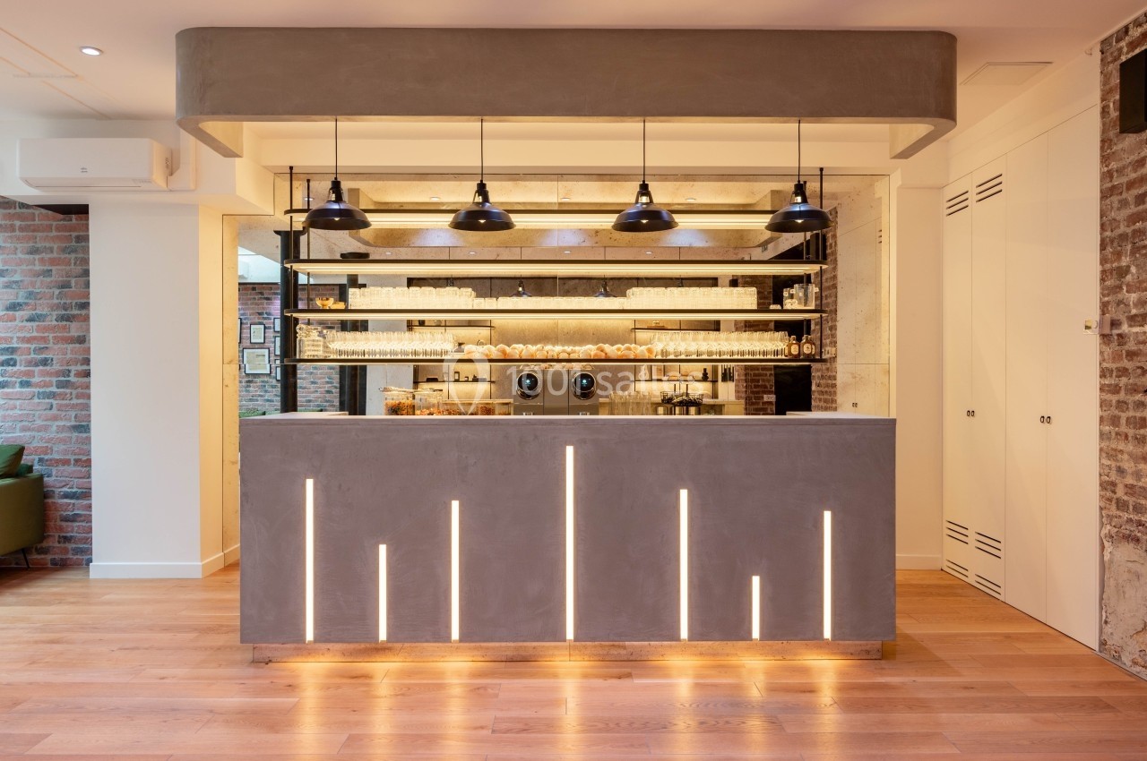 Comptoir moderne en béton éclairé, avec étagères remplies de tasses et lampes suspendues dans un intérieur lumineux.