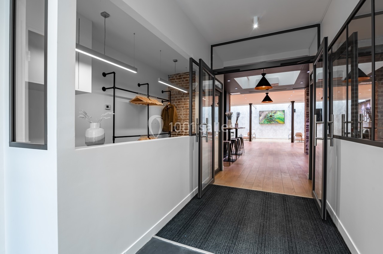 Entrée d'un espace moderne avec murs blancs, sol en bois, tapis gris et portes vitrées ouvertes sur une salle lumineuse.