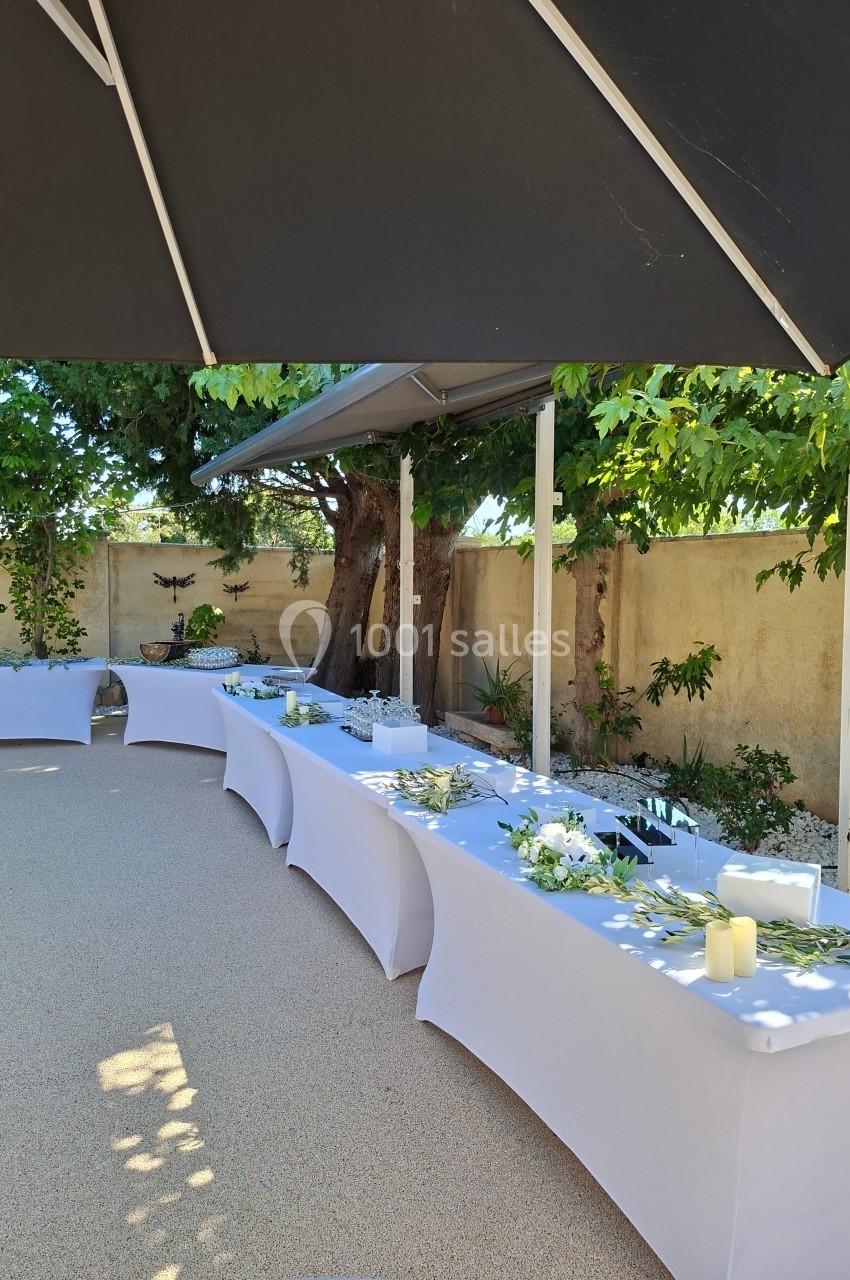 Tables décorées avec nappes blanches et ornements floraux, disposées en extérieur sous un parasol et des arbres.