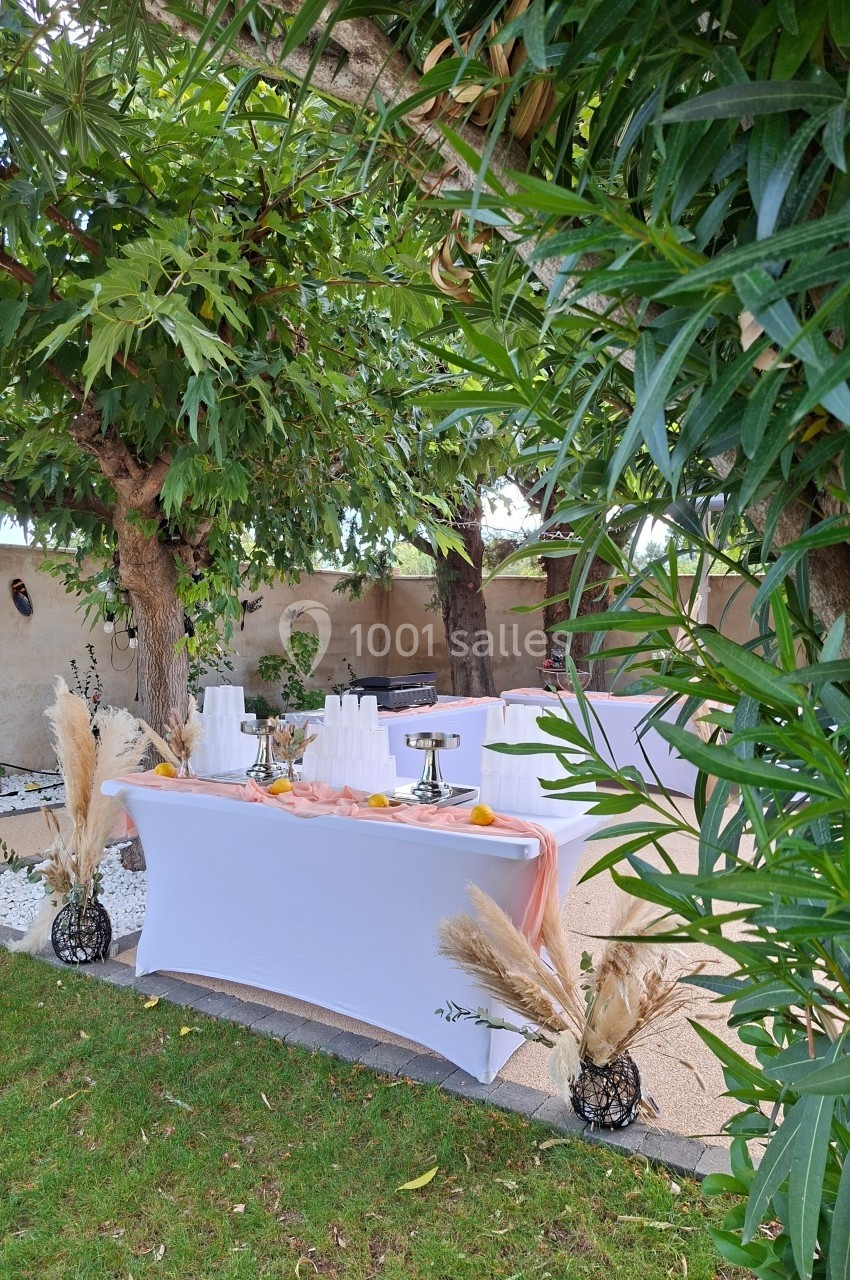Table dressée en extérieur sous des arbres, décorée de nappes blanches, pampas et vaisselle pour un événement.