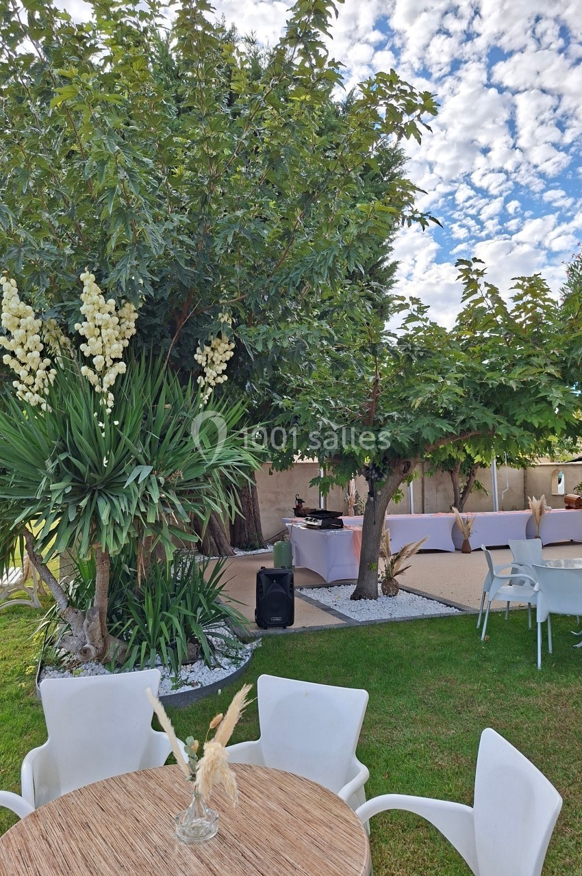 Espace extérieur avec tables, chaises blanches, arbres et plantes, décoré pour un événement sous un ciel partiellement…
