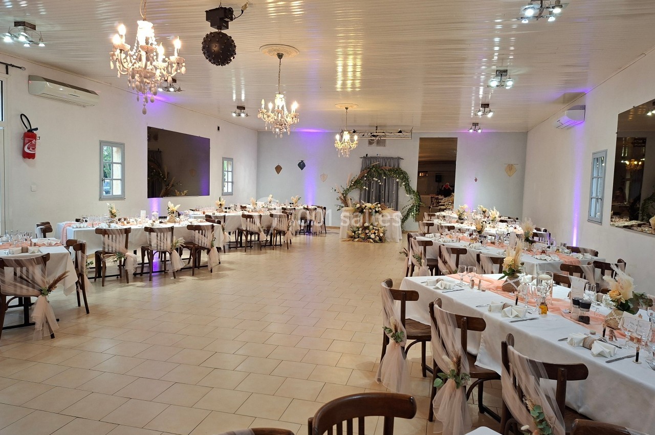 Salle de réception décorée pour un événement, avec tables dressées, chaises ornées et éclairage chaleureux.