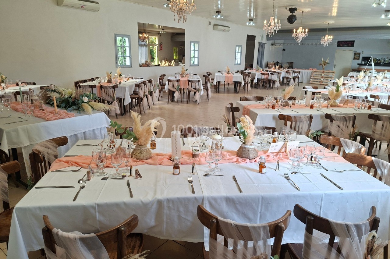 Salle de réception décorée pour un événement, avec des tables dressées, nappes blanches et chemins de table roses.