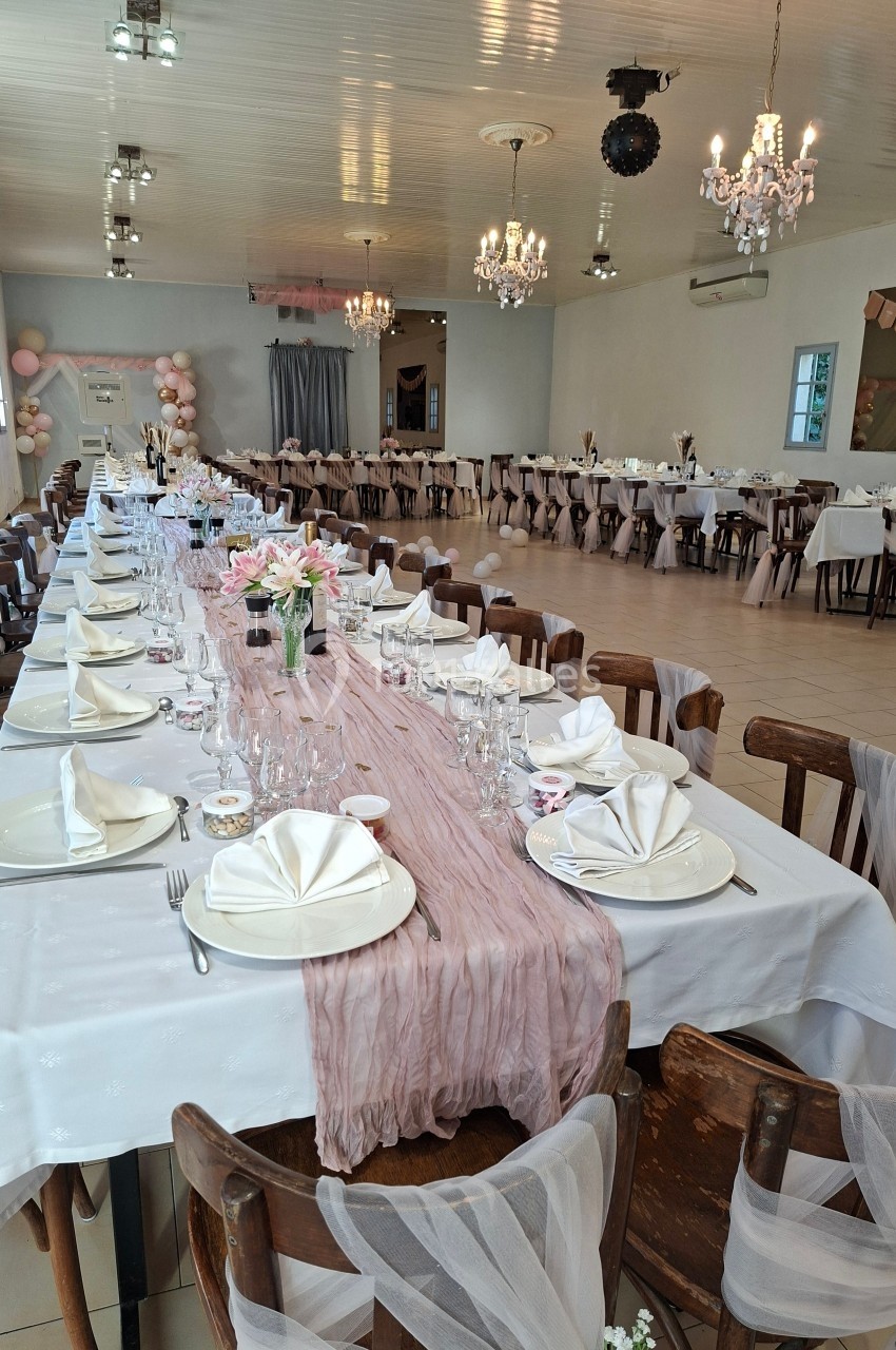Salle de réception décorée avec des tables alignées, nappes blanches, chemins de table roses et chaises ornées de tissu.