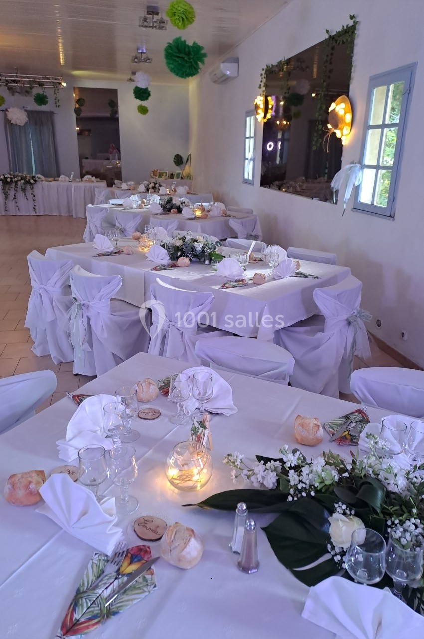 Salle décorée pour un événement, avec des tables dressées, nappes blanches, centres de table floraux et éclairage doux.