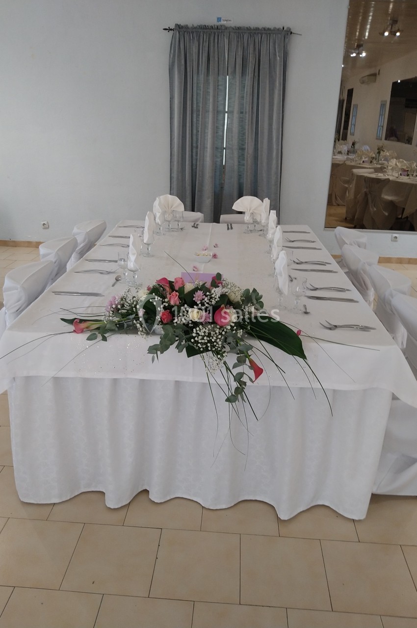 Table rectangulaire décorée pour un événement, avec nappes blanches, couverts alignés et centre floral coloré.