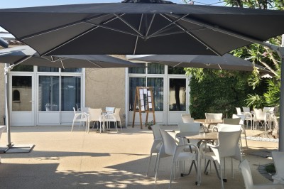Terrasse extérieure avec tables et chaises blanches, ombragée par des parasols noirs et entourée de végétation.