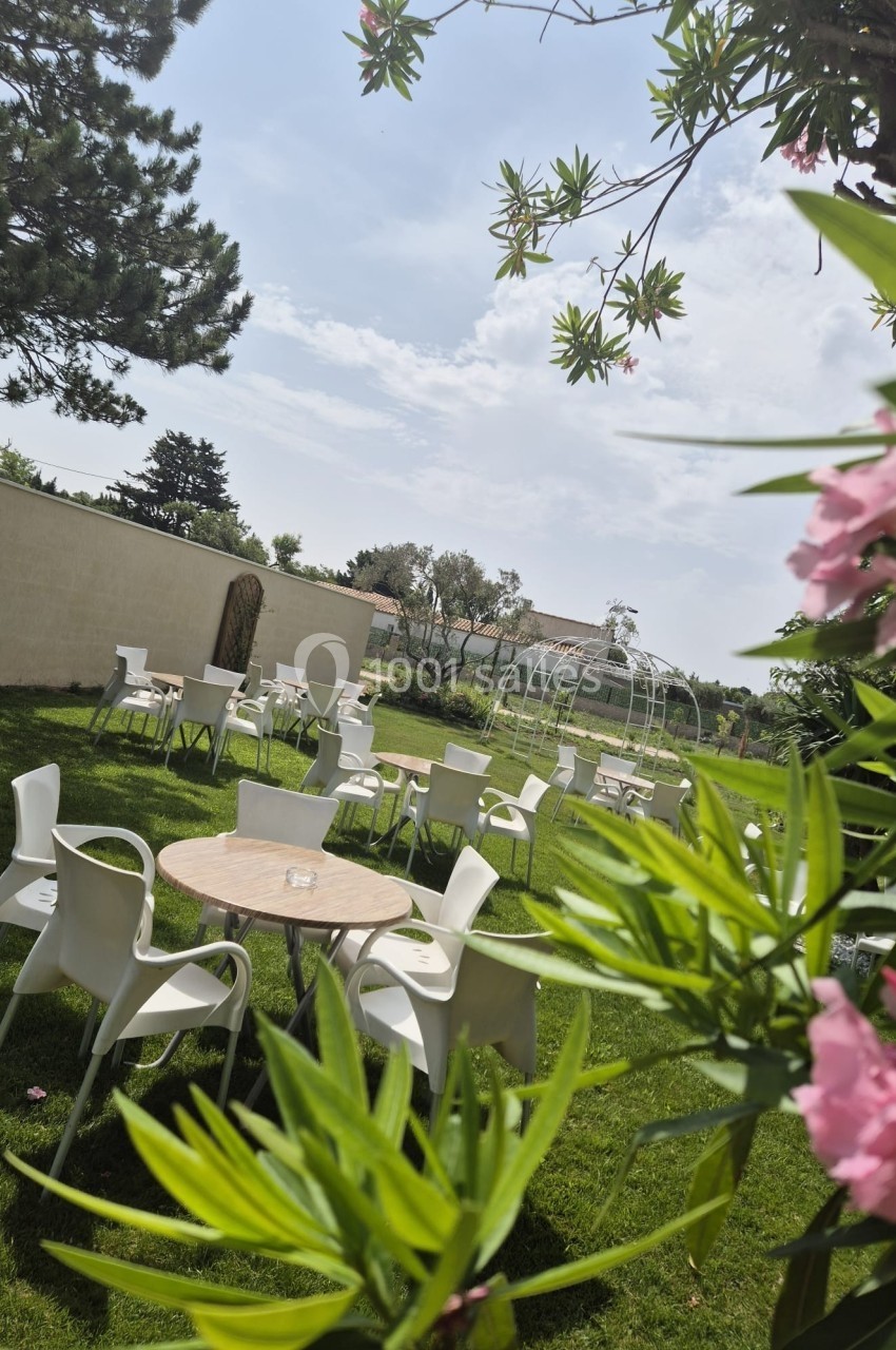 Chaises et tables disposées dans un jardin verdoyant, entourées de fleurs et d'arbres sous un ciel clair.