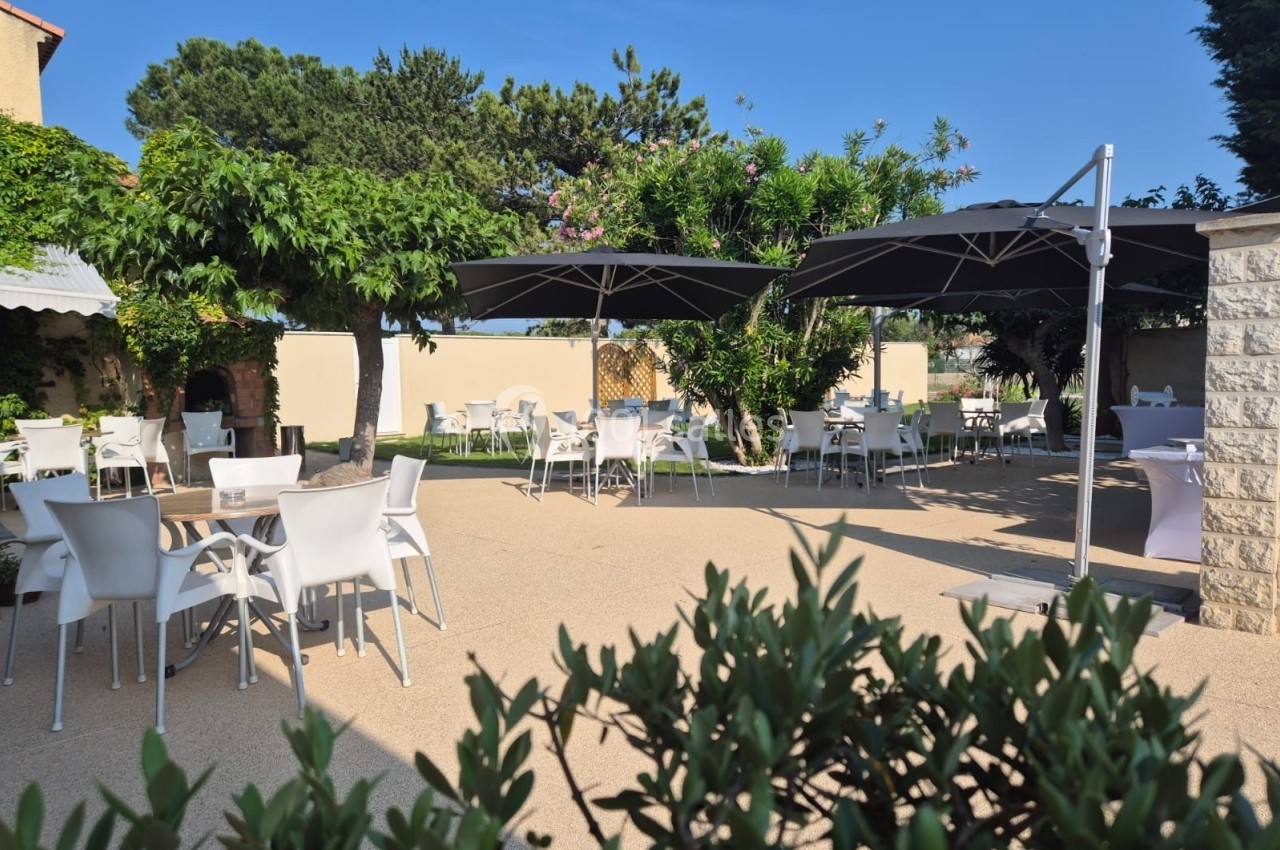 Terrasse extérieure avec tables et chaises blanches, ombragée par des parasols noirs et entourée de végétation.