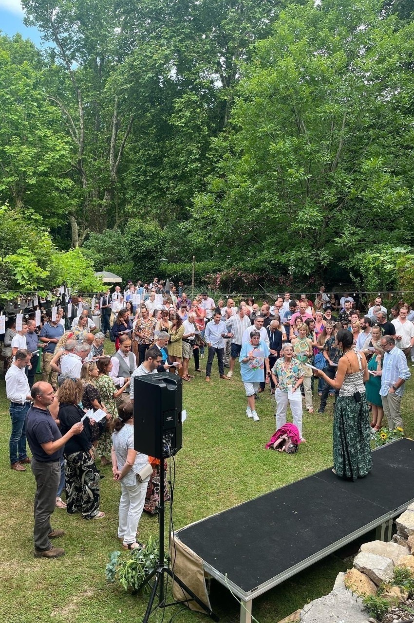 Un groupe de personnes rassemblées en plein air près d'une scène, entourées de verdure et d'arbres.