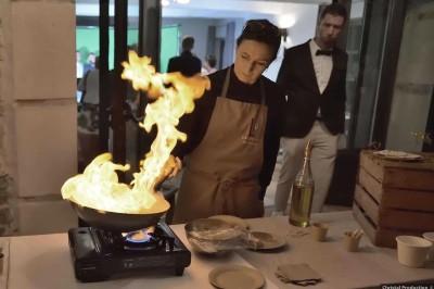 Bouteilles en verre suspendues contenant des fleurs et des branches, devant une maison en pierre entourée de verdure.