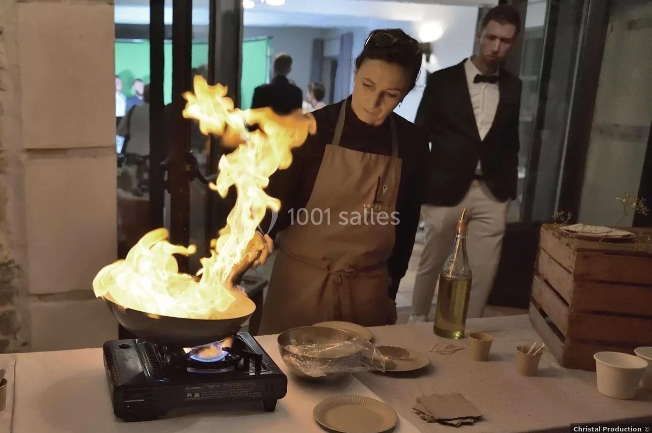 Une personne en tablier cuisine devant une poêle enflammée sur un réchaud, entourée de vaisselle et d'ingrédients.