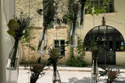 Bouteilles en verre suspendues contenant des fleurs et des branches, devant une maison en pierre entourée de verdure.