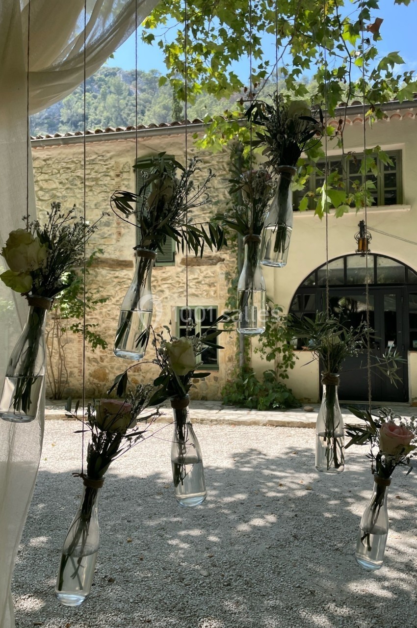 Bouteilles en verre suspendues contenant des fleurs et des branches, devant une maison en pierre entourée de verdure.
