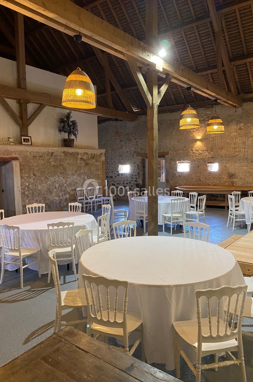 Salle rustique avec murs en pierre, tables rondes dressées de nappes blanches et chaises en bois, éclairée par des…