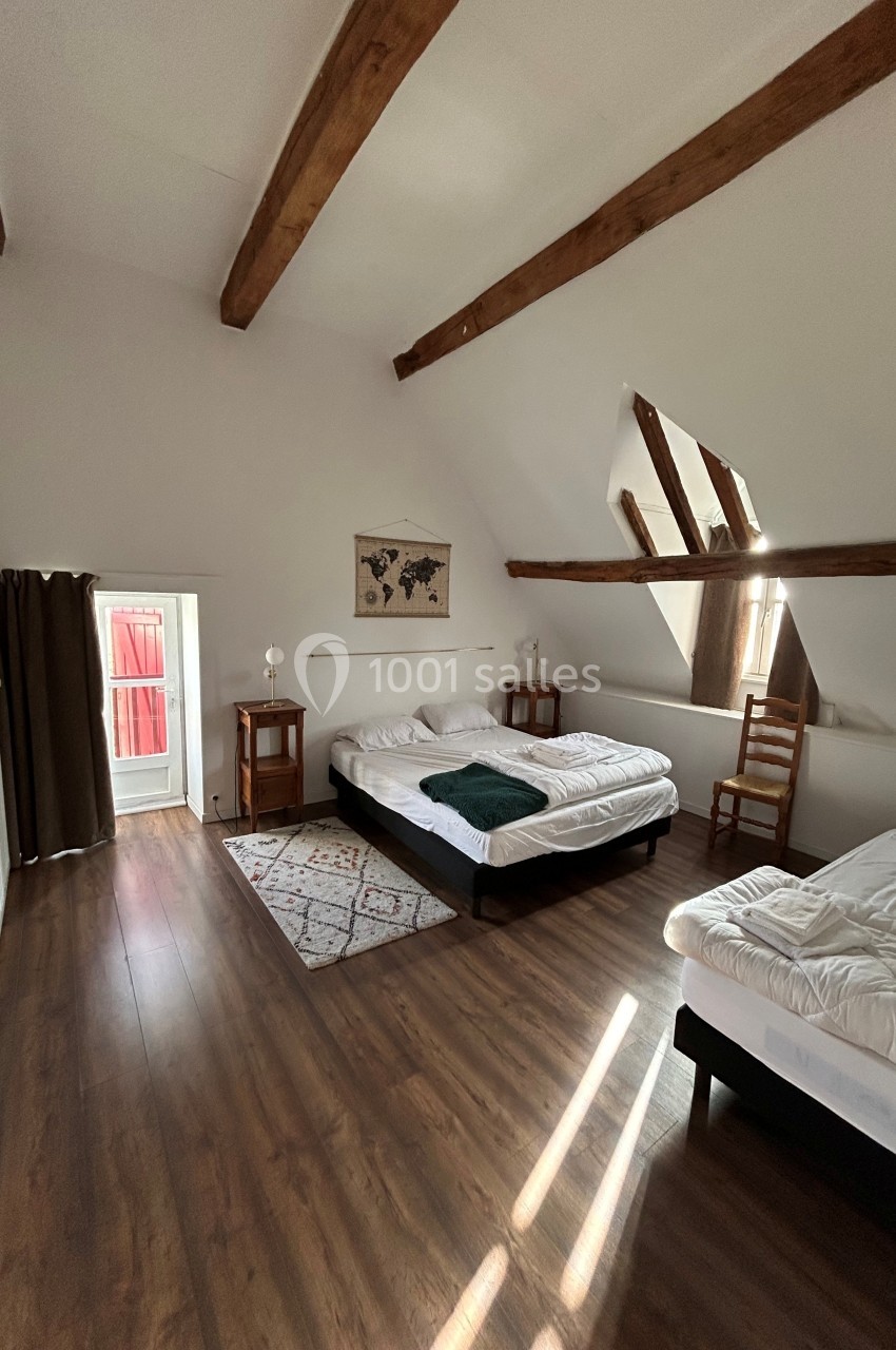 Chambre lumineuse avec parquet, poutres apparentes, deux lits doubles, tapis, chaises et petite fenêtre donnant sur l…