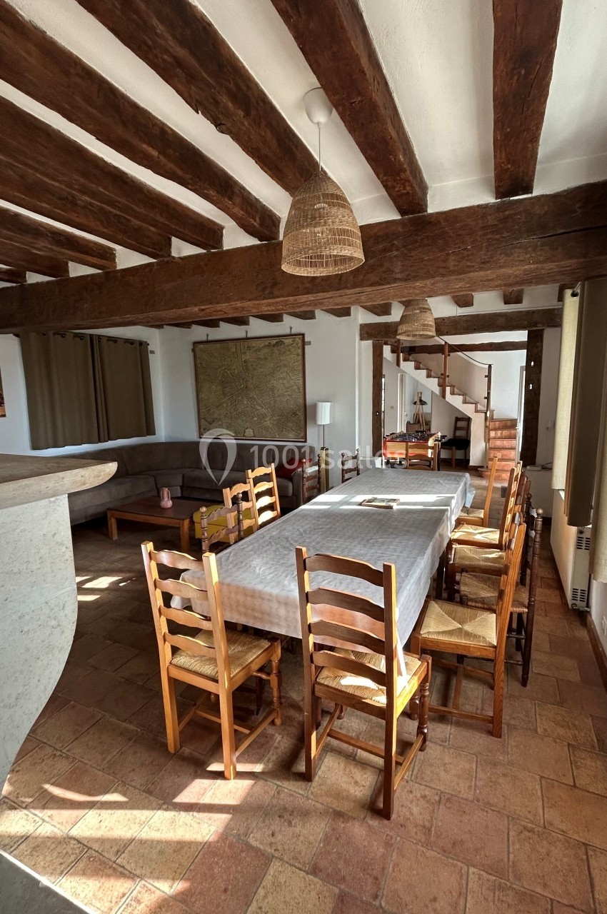 Salle à manger rustique avec une grande table rectangulaire entourée de chaises en bois, poutres apparentes au plafond.