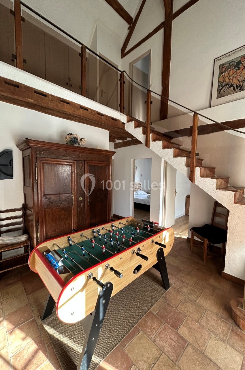Table de baby-foot en bois dans une pièce lumineuse avec sol en carrelage, escalier en bois et mezzanine.