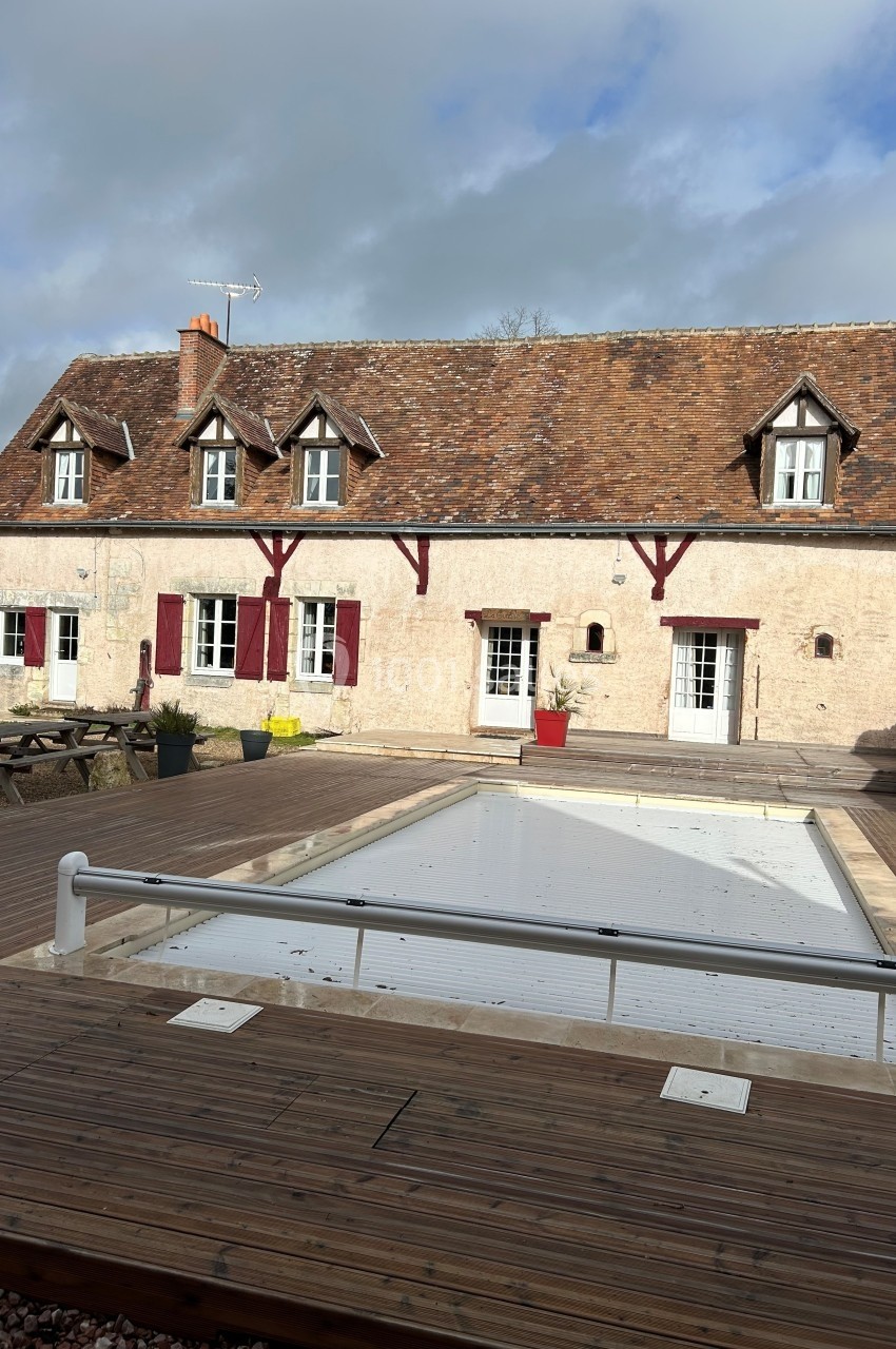 Maison en pierre avec toit en tuiles, volets rouges et piscine couverte sur une terrasse en bois.