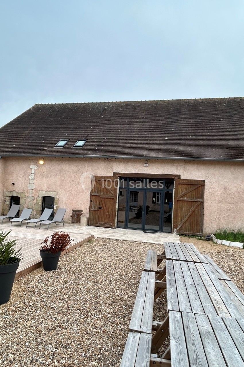 Façade d'une maison en pierre avec portes en bois ouvertes, terrasse en gravier et table de pique-nique en premier plan.