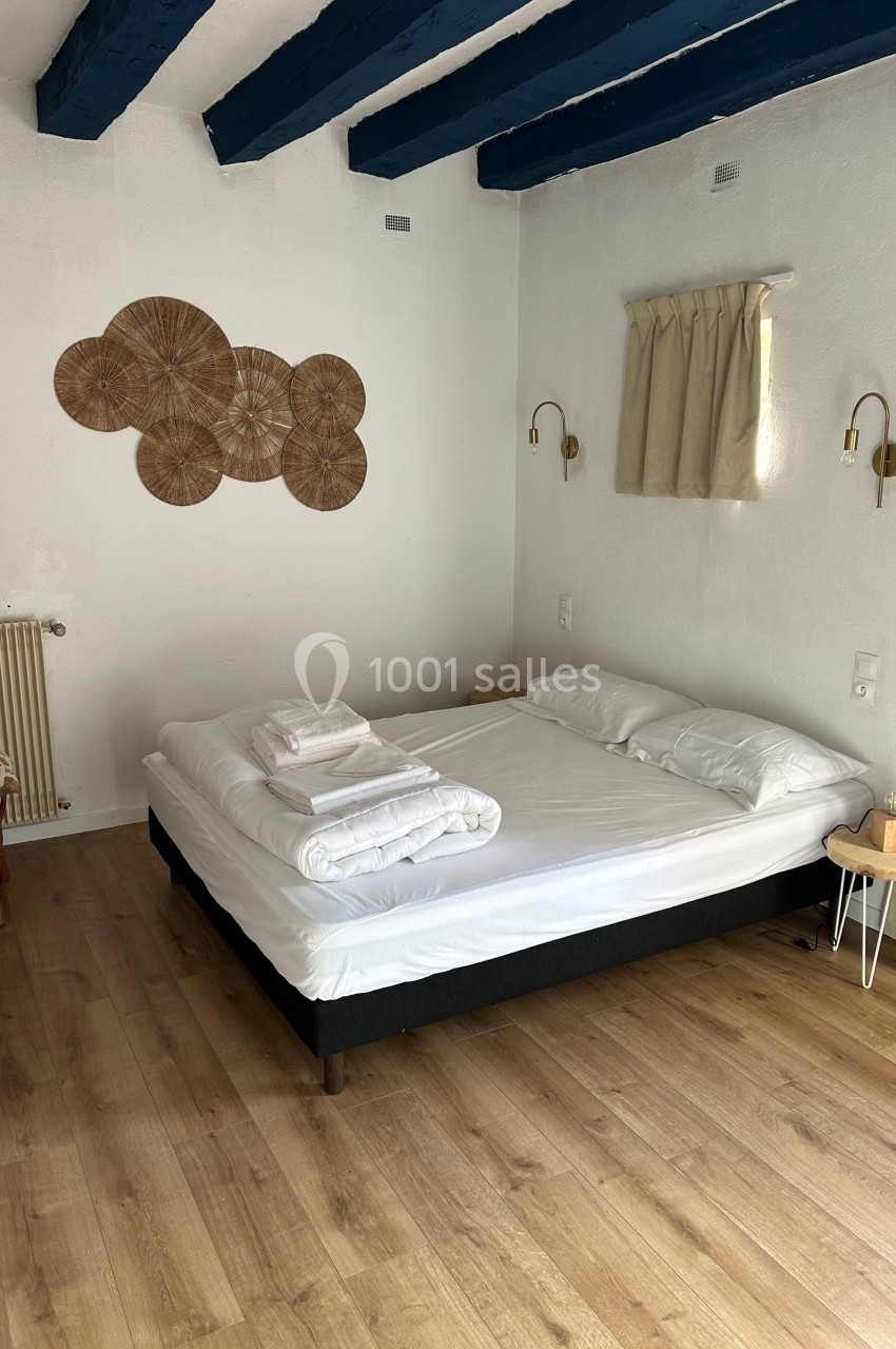 Chambre lumineuse avec un lit double, draps pliés, parquet en bois, murs blancs et poutres apparentes bleues.