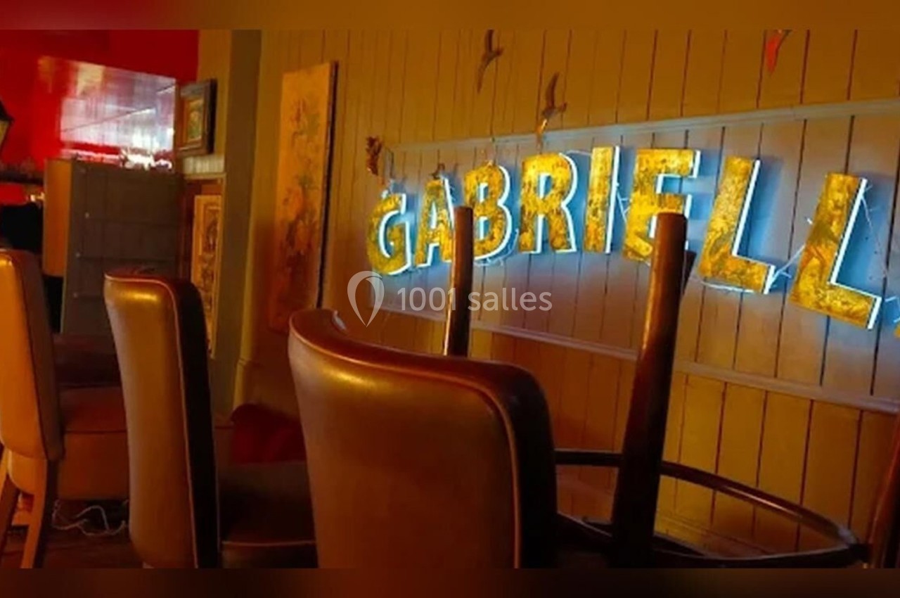 Chaises hautes retournées sur un comptoir dans un espace intérieur avec une enseigne lumineuse ’Gabriella’ au mur.