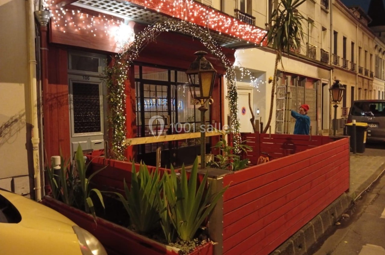 Terrasse d'un café décorée de guirlandes lumineuses, entourée de plantes, dans une rue calme en soirée.