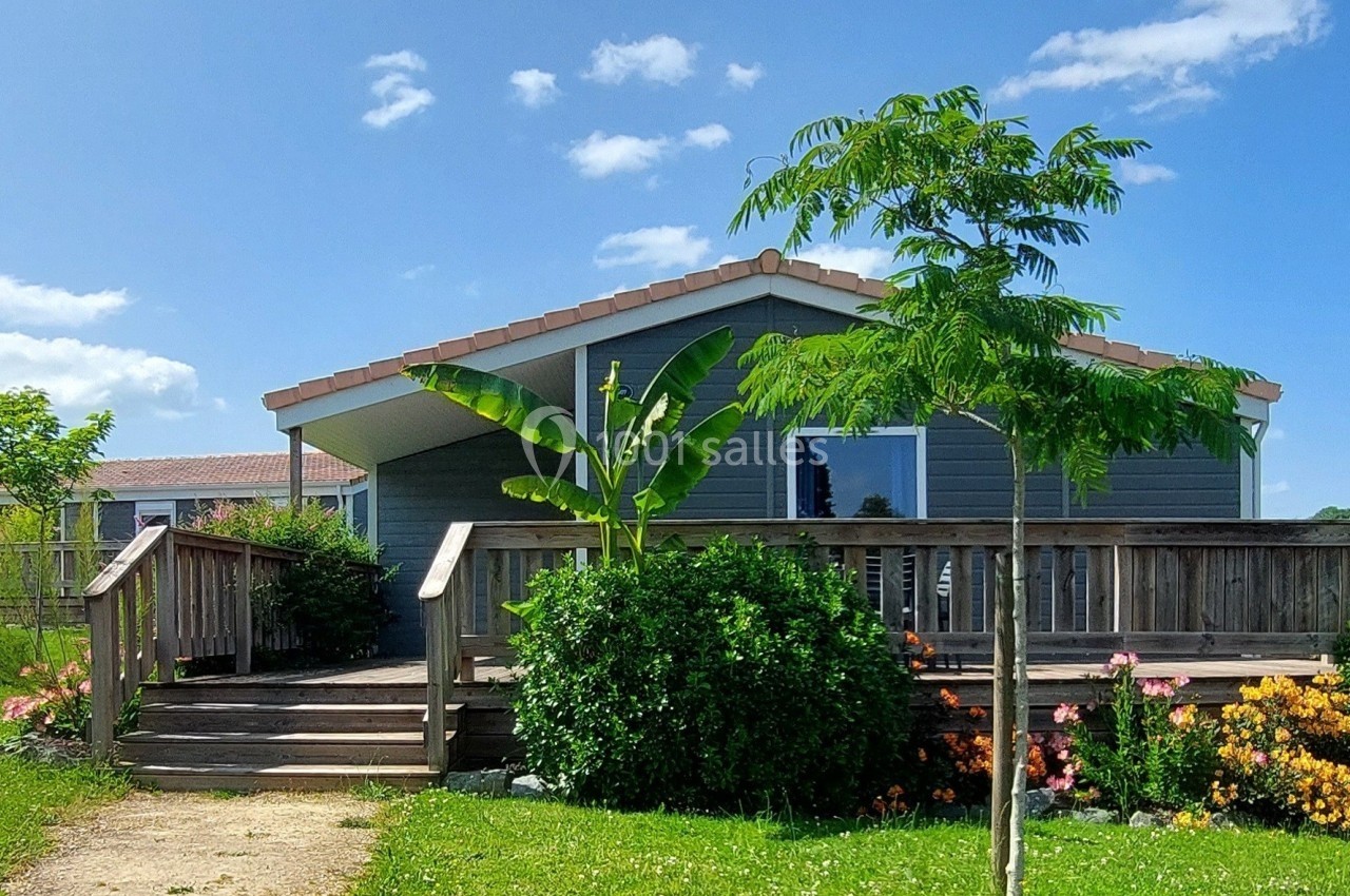 Maison en bois avec terrasse entourée de plantes et d'arbres, sous un ciel bleu clair.