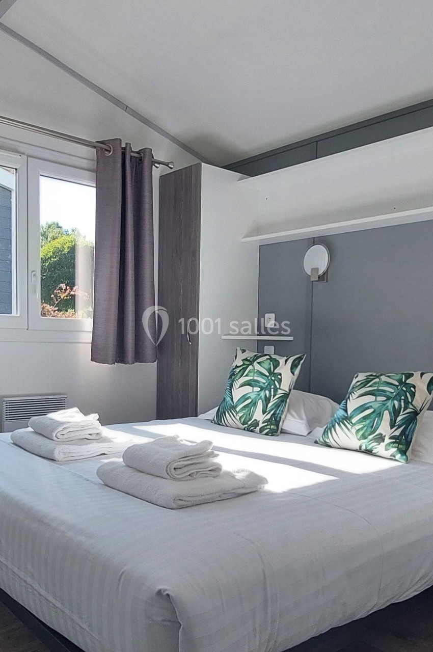 Chambre lumineuse avec lit double, draps blancs, coussins à motifs végétaux, fenêtres avec rideaux et éclairage mural.